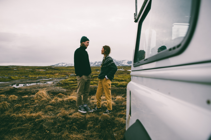 Een man en vrouw staan naast een huurauto in Scandinavië, met bergen op de achtergrond.